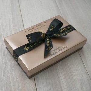A closed box of our eight piece dark assortment. The lid of the box is a light brown and has a black bow on it that reads John Kelly Chocolates on it.